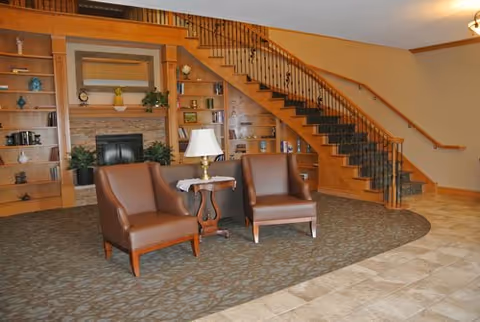 A cozy living area with two brown leather chairs and a small wooden table with a lamp between them. Behind the chairs is a built-in wooden bookshelf with decorative items and a fireplace in the center. A staircase with wooden steps and black iron railings curves upward on the right side of the image. The floor is a combination of carpet and tile.