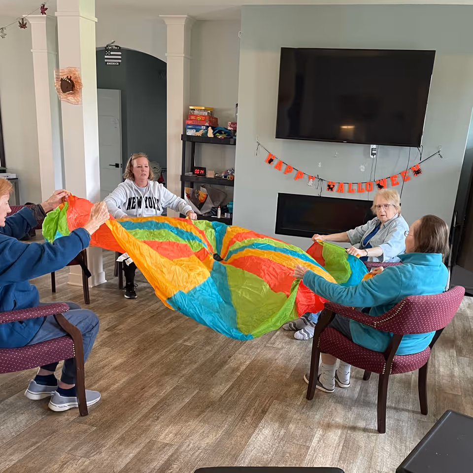 Four elderly women sitting in a circle in a living room, holding the edges of a colorful parachute-like fabric. The room has a large flat-screen TV mounted above a fireplace, and a 'Happy Halloween' banner is hanging on the wall. There are shelves with board games and other items in the background.