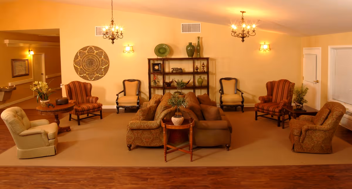 A warm and inviting living room area in an assisted living facility with multiple upholstered armchairs and sofas arranged around small wooden tables. The walls are painted a soft yellow and decorated with a large circular wall art piece and a wooden shelving unit holding decorative vases and plants. Two chandeliers and wall sconces provide lighting, and there is a window with closed blinds on the right side.