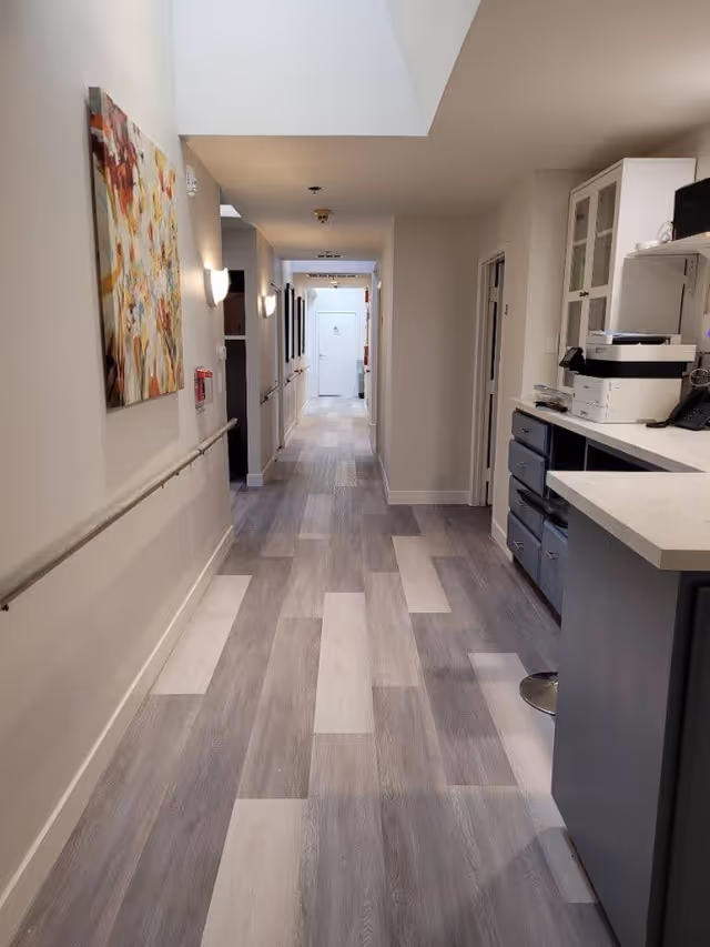 A long, well-lit hallway with light-colored wood flooring and beige walls. On the left wall, there is a colorful abstract painting and a handrail running along the length of the hallway. On the right side, there is a counter with office equipment including a printer and telephone. The hallway leads to a white door at the far end.