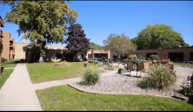 Outdoor courtyard area of a senior living community with green grass, trees, flower beds, and several benches and tables arranged on a paved surface under a clear blue sky.