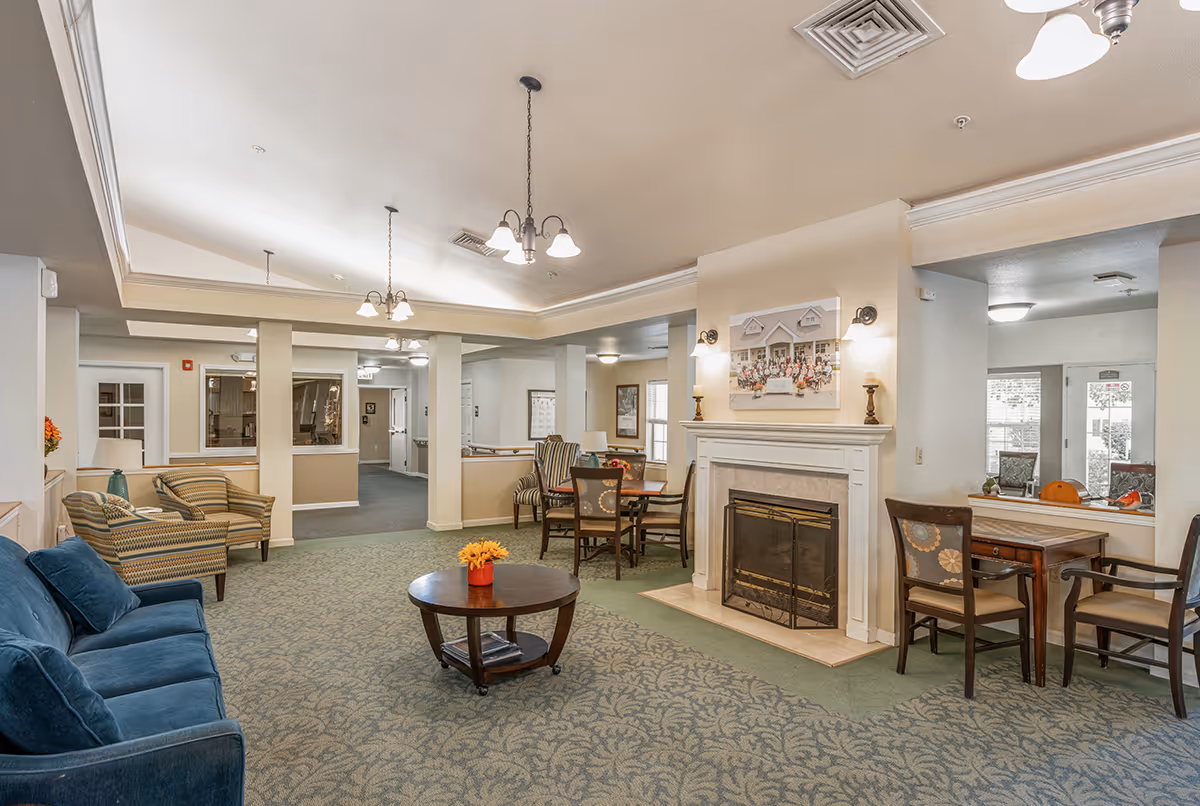 Spacious senior living common room with sofas and chairs, a central coffee table, dining tables, and a fireplace under hanging lights.
