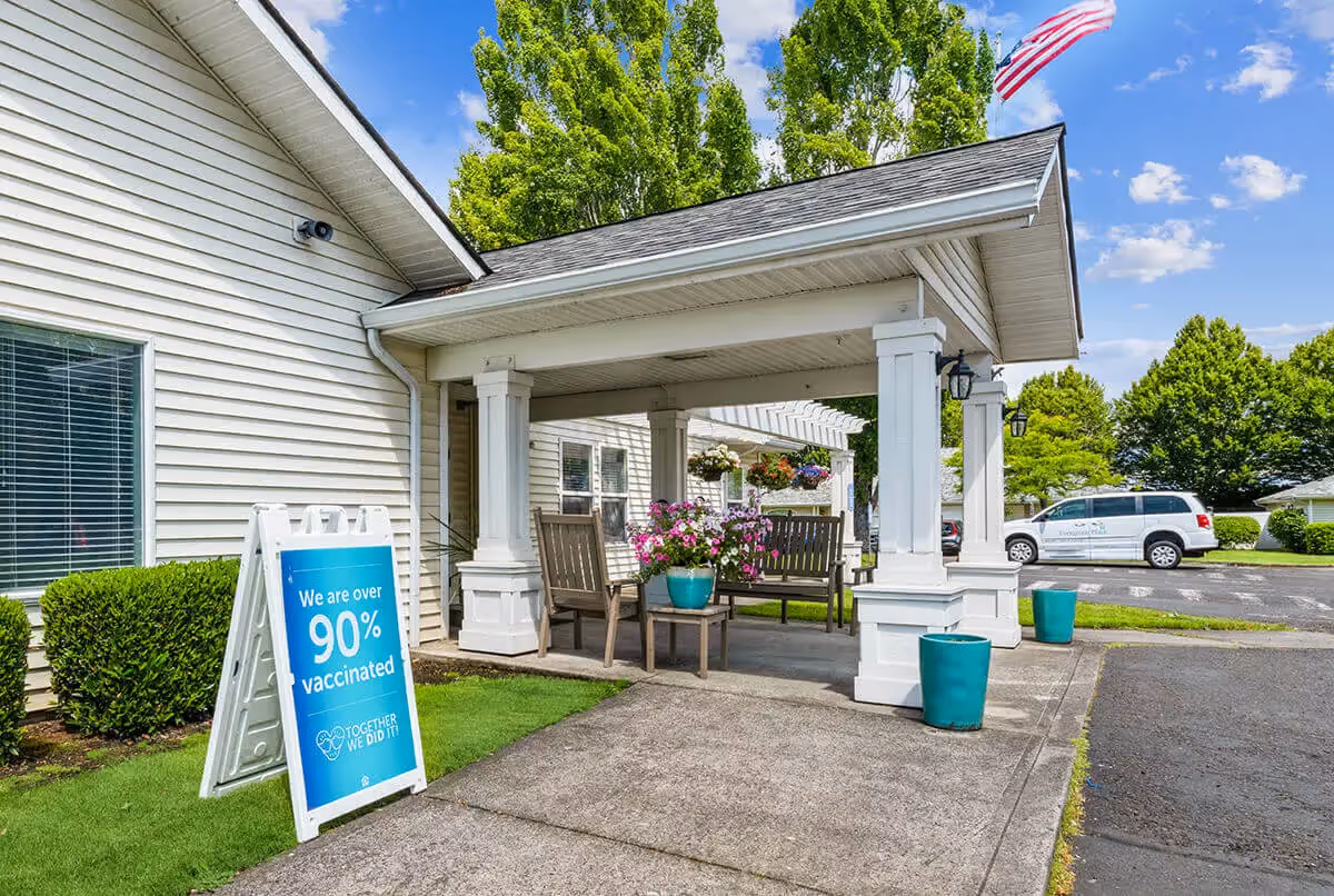 Outdoor covered seating area at Evergreen Place with wooden benches and a small table holding a pot of pink flowers. A sign nearby reads 'We are over 90% vaccinated TOGETHER WE DID IT!'. There are green plants and trees around, a parked white van, and an American flag flying in the background under a blue sky with some clouds.