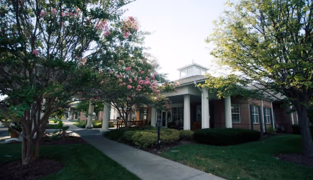 Front entrance of a single-story senior living building with a covered porch, columns, flowering trees and a sidewalk.