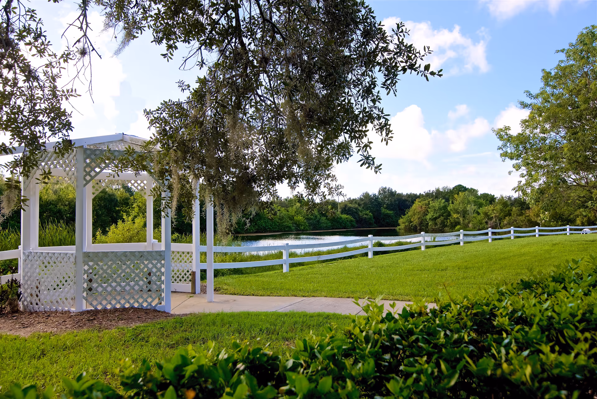 A white wooden gazebo with lattice sides situated on a grassy area near a pond, surrounded by green trees and bushes under a partly cloudy blue sky.