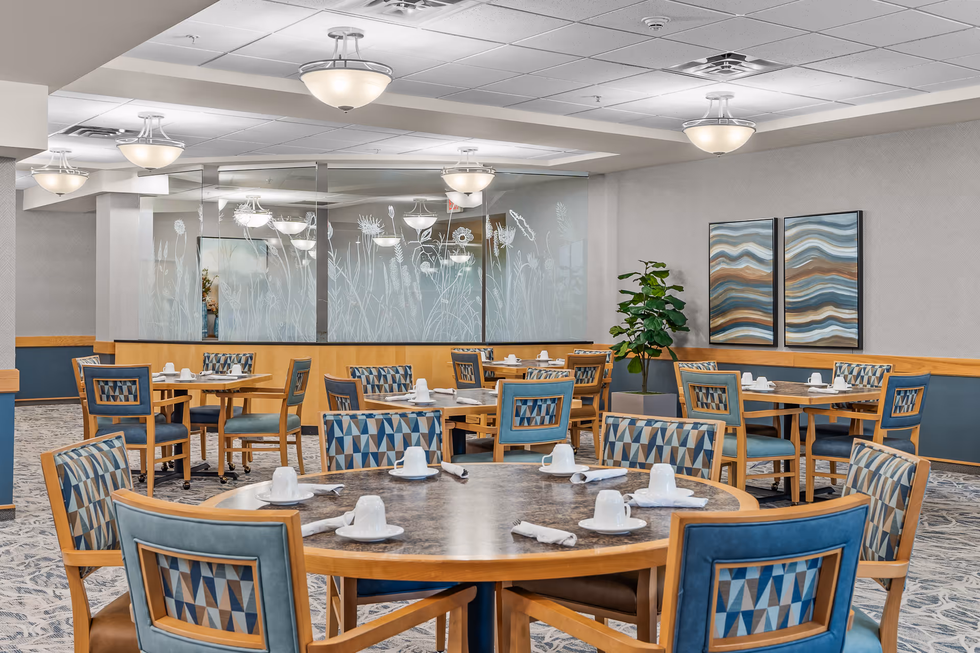 Bright dining room with round and rectangular tables set with cups and napkins, patterned chairs, decorative glass partition, artwork, and ceiling lights.