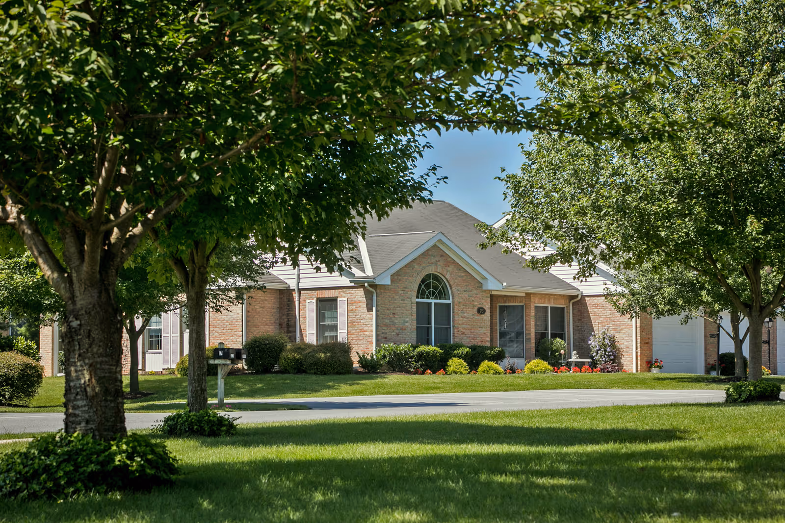 A single-story brick residential building with a well-maintained lawn and several trees in the foreground under a clear blue sky.