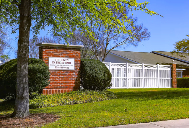 Outdoor view of The Haven in the Summit facility sign made of red brick with white text, surrounded by green bushes and grass, with a white fence and building in the background under a clear blue sky.