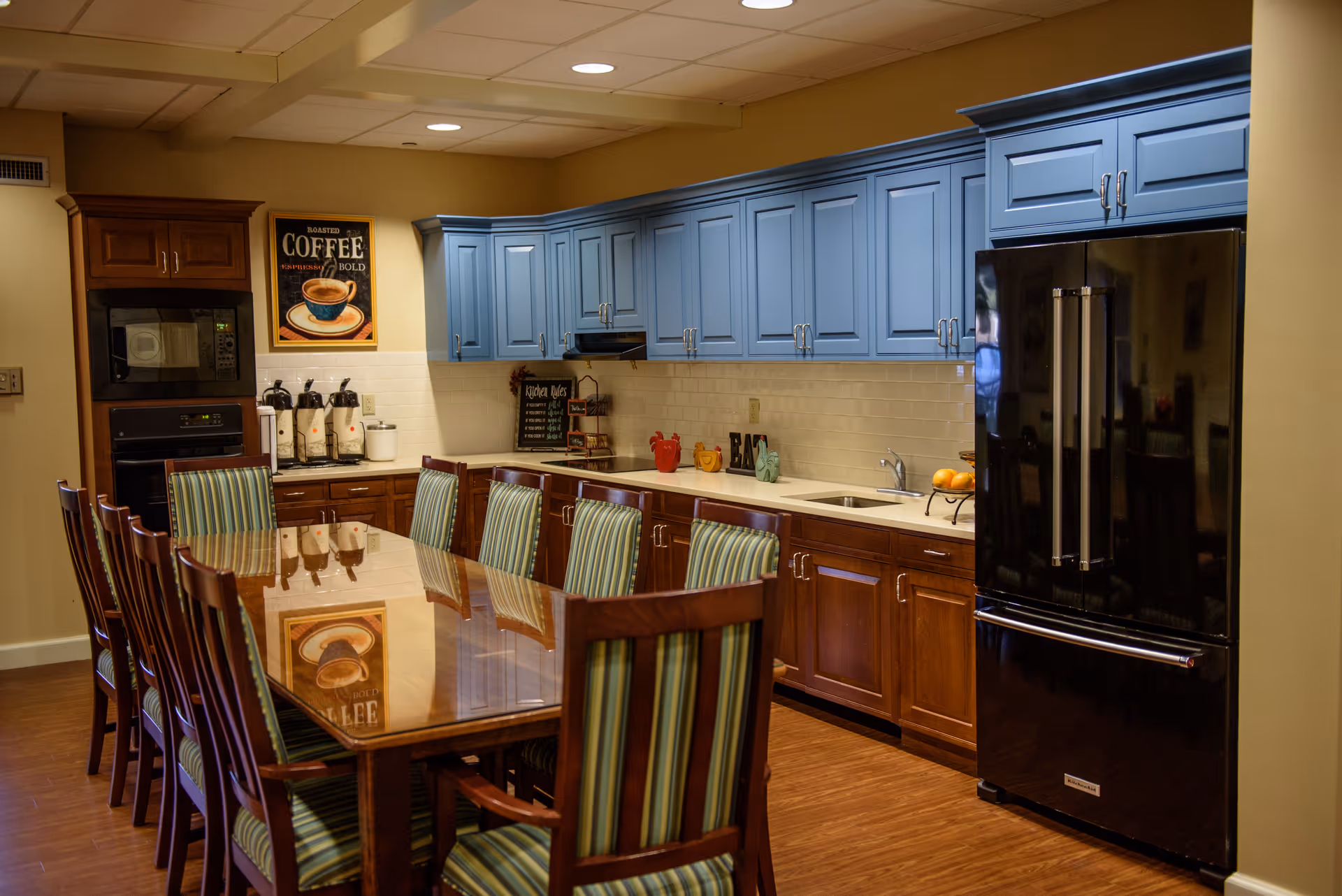 Communal kitchen and dining area with a long wooden table and striped chairs, blue upper cabinets, wooden lower cabinets and a black refrigerator.