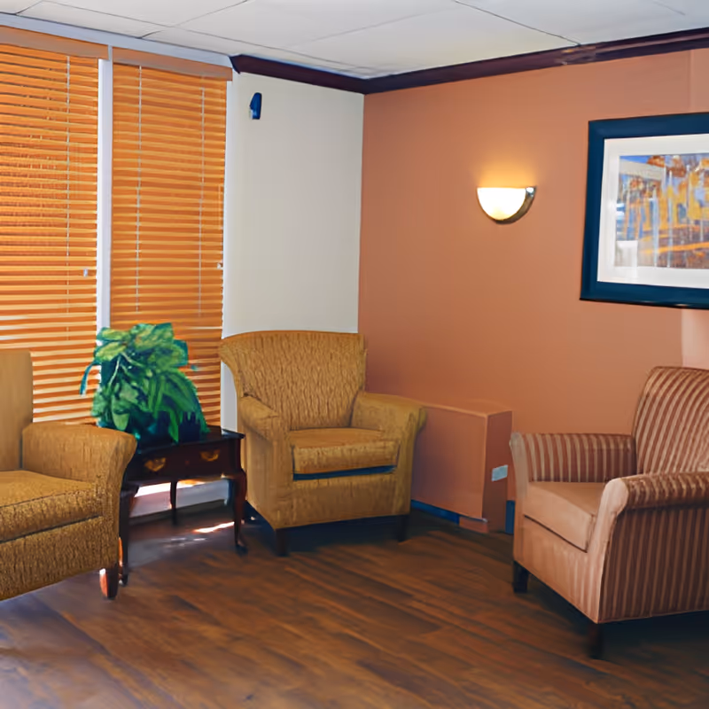 A cozy sitting area with three upholstered armchairs arranged around a small wooden side table with a green potted plant. The room has wooden blinds covering the windows, a warm-toned accent wall with a wall-mounted light fixture, and a framed abstract painting.