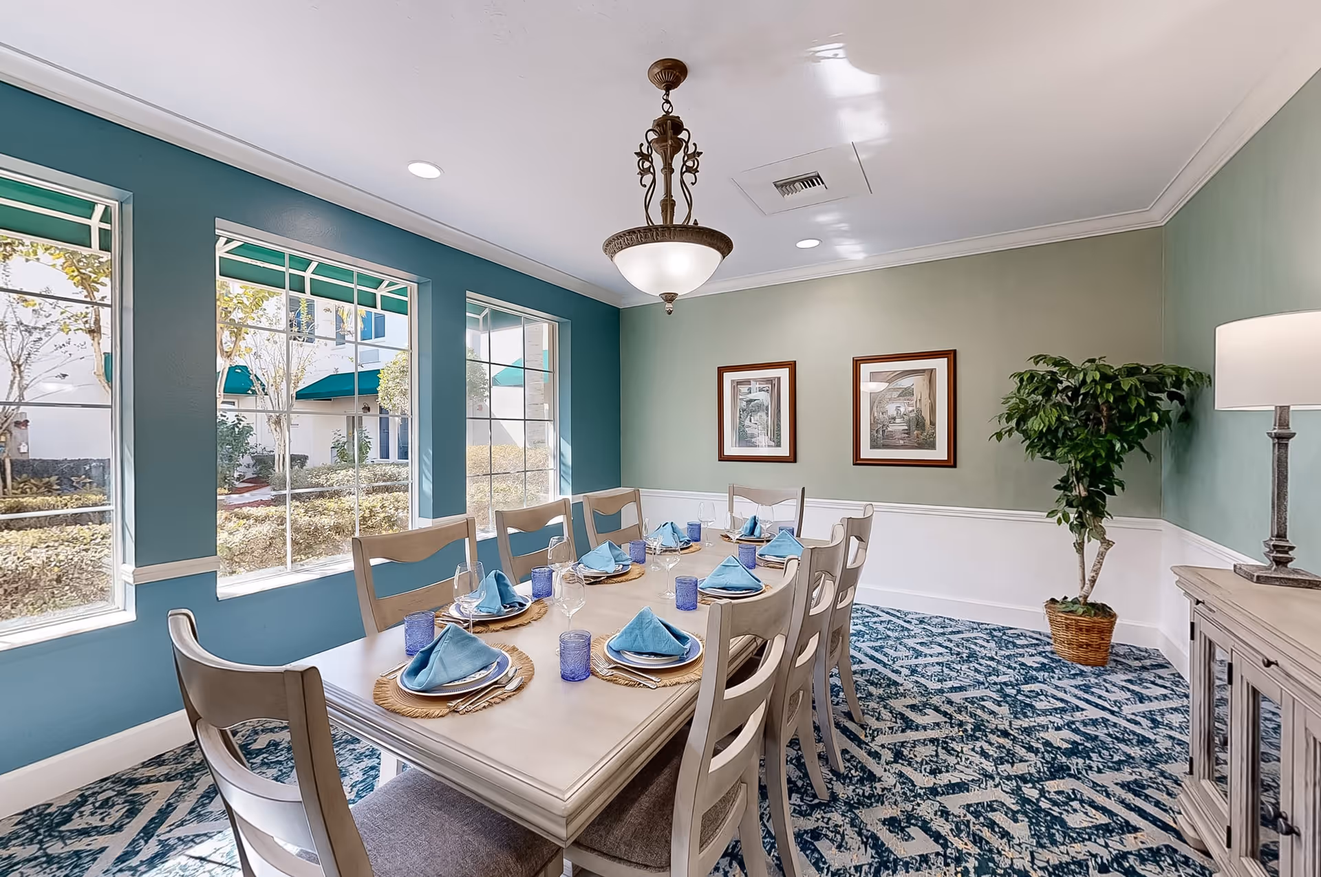 A bright dining room with a long rectangular table set for eight people. The table is adorned with blue napkins, plates, and blue drinking glasses. The room has large windows with green awnings outside, allowing natural light to fill the space. The walls are painted in shades of green and teal, decorated with two framed pictures and a potted plant in the corner. A chandelier hangs above the table, and a lamp is placed on a sideboard against the wall.