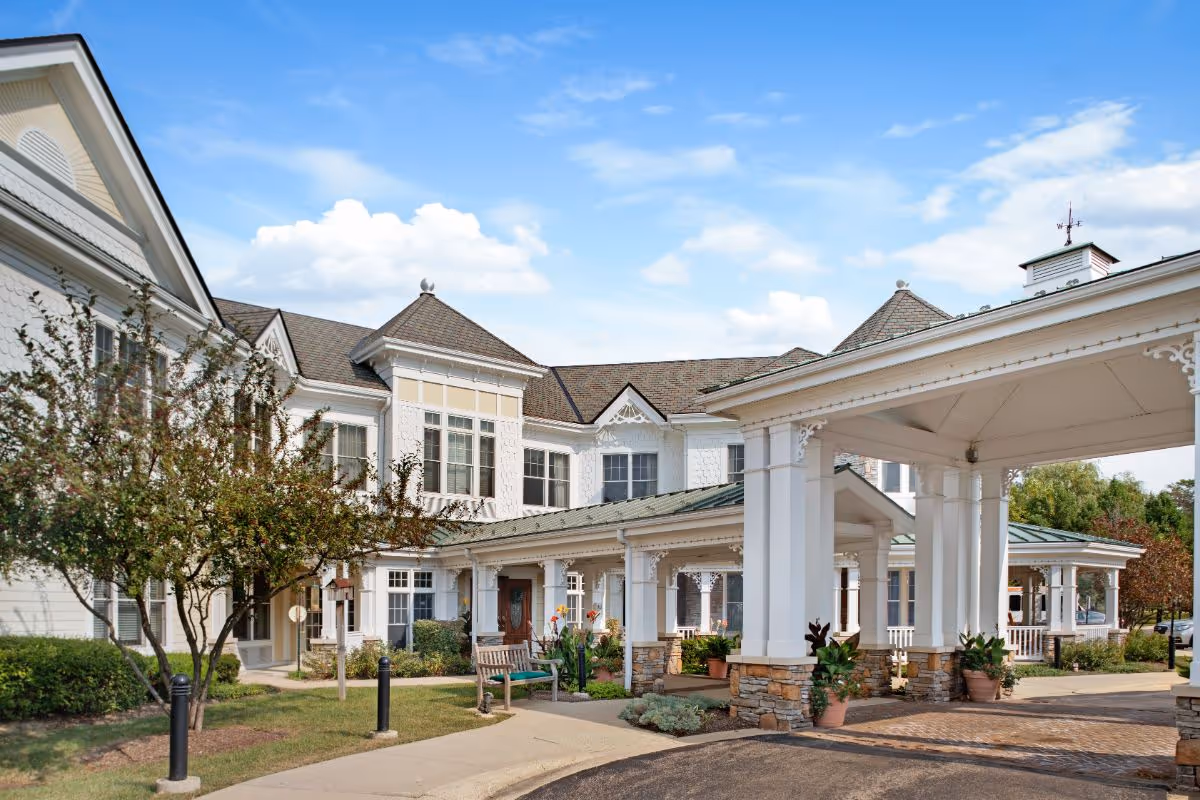 Exterior view of a senior living facility with white siding and stone accents, featuring a covered entrance with columns, landscaped greenery, and a bench near the walkway under a partly cloudy blue sky.