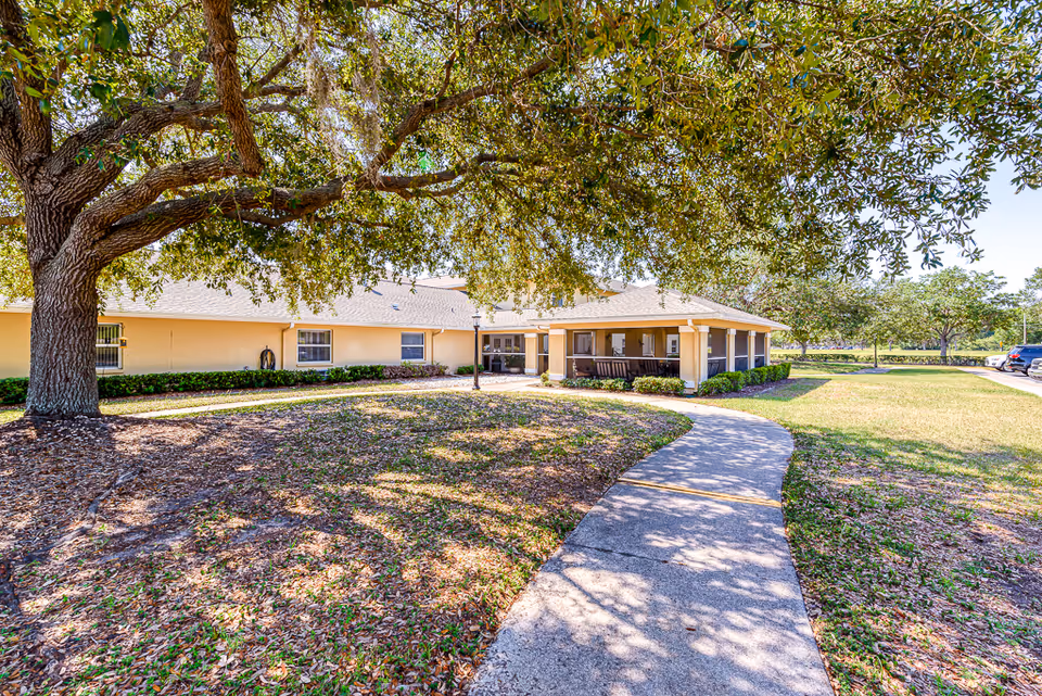 A sunny outdoor view of a senior living facility with a large tree casting shadows over a curved concrete walkway leading to a covered porch area of a single-story building. The building is light yellow with several windows and surrounded by grass and small bushes. A few cars are parked in the distance on the right side.