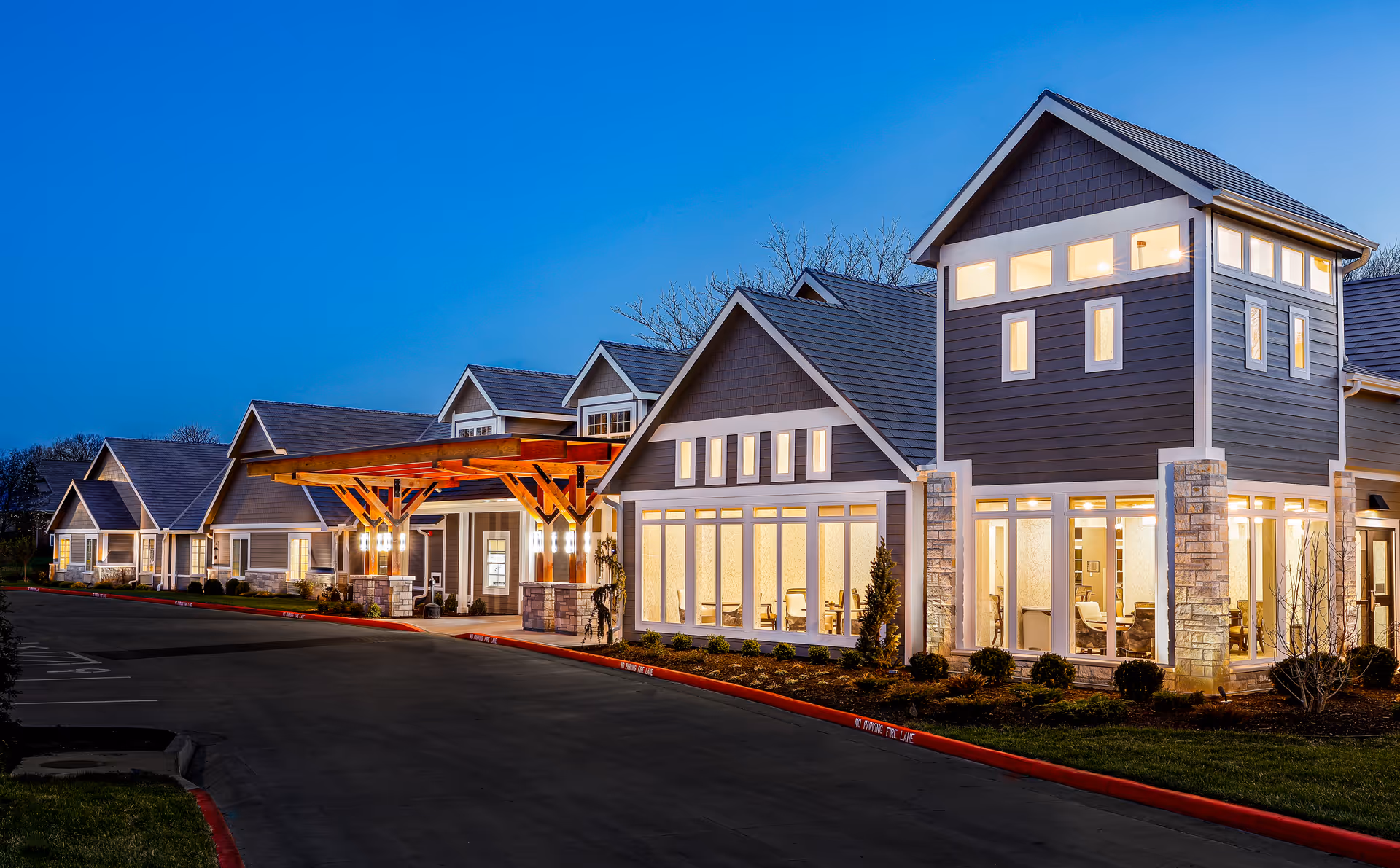 Exterior view of Homestead Estates Assisted Living of Leawood at dusk, showing a modern building with multiple peaked roofs, large windows illuminated from inside, and a covered entrance with wooden beams. The surrounding area includes a driveway and landscaped greenery.