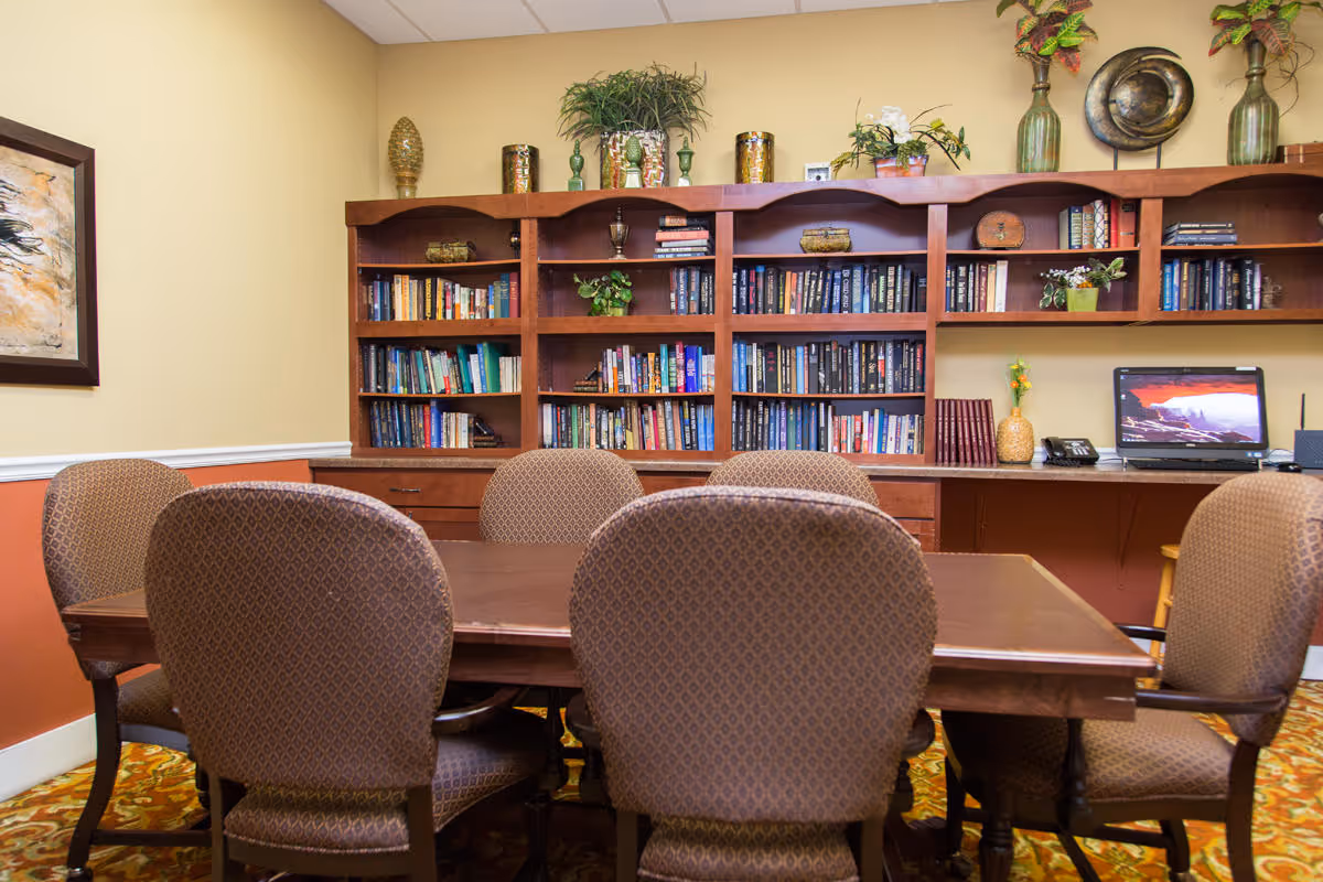 A cozy room with a wooden table surrounded by six upholstered chairs. Behind the table is a large wooden bookshelf filled with books and decorative items. On the right side of the bookshelf, there is a desk with a computer, a phone, and some plants. The walls are painted beige and orange, and the floor has a patterned carpet.