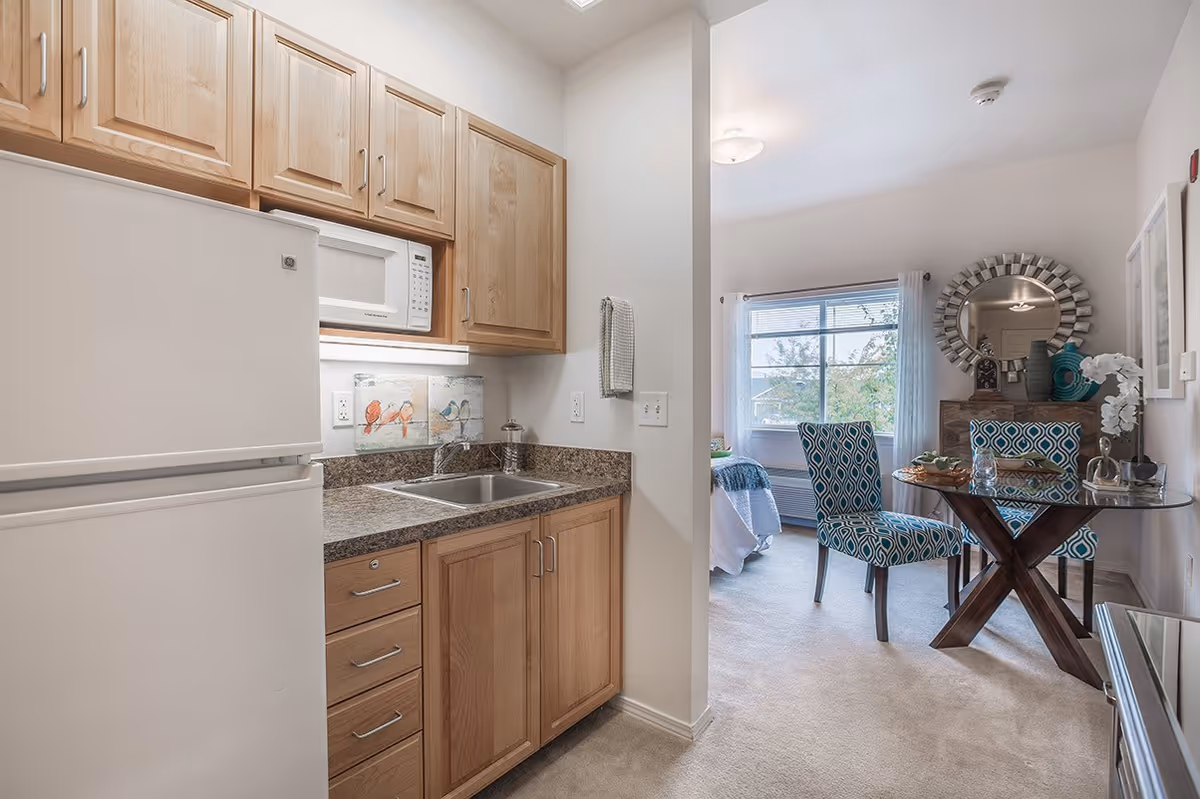 A small kitchen area with wooden cabinets, a white refrigerator, a microwave, and a sink with a granite countertop. Adjacent to the kitchen is a dining area with a glass-top table and two patterned blue chairs. A large window with white curtains lets in natural light, and a decorative round mirror hangs on the wall above a wooden sideboard.