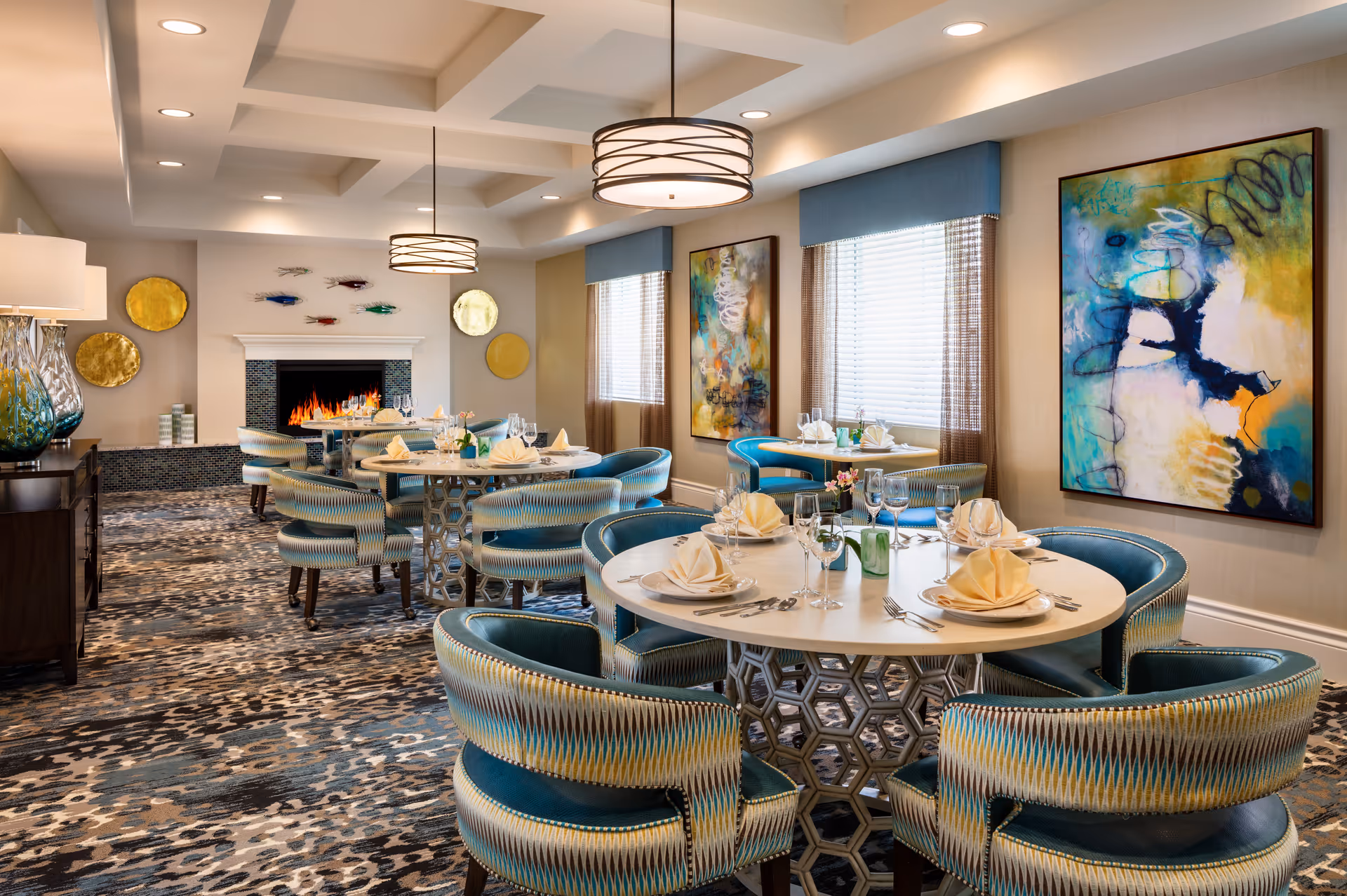 Elegant dining room with round tables set for meals, teal upholstered chairs, a fireplace, and colorful wall art.