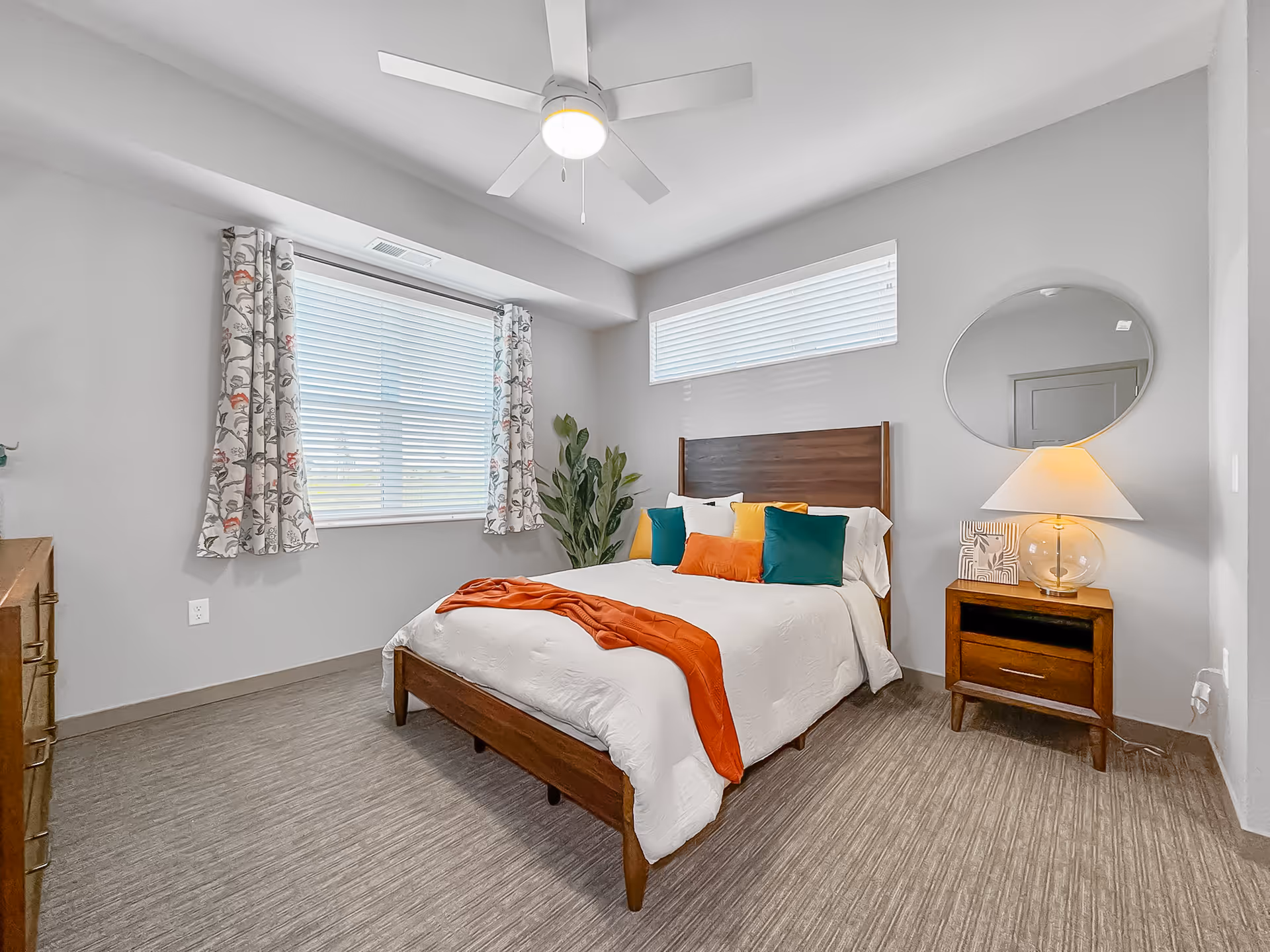 A bright bedroom with a wooden bed frame and white bedding accented by colorful pillows and an orange throw blanket. The room features a ceiling fan with a light, a large window with floral curtains, a smaller horizontal window above the bed, a wooden nightstand with a lamp and decorative items, and a round mirror on the wall.