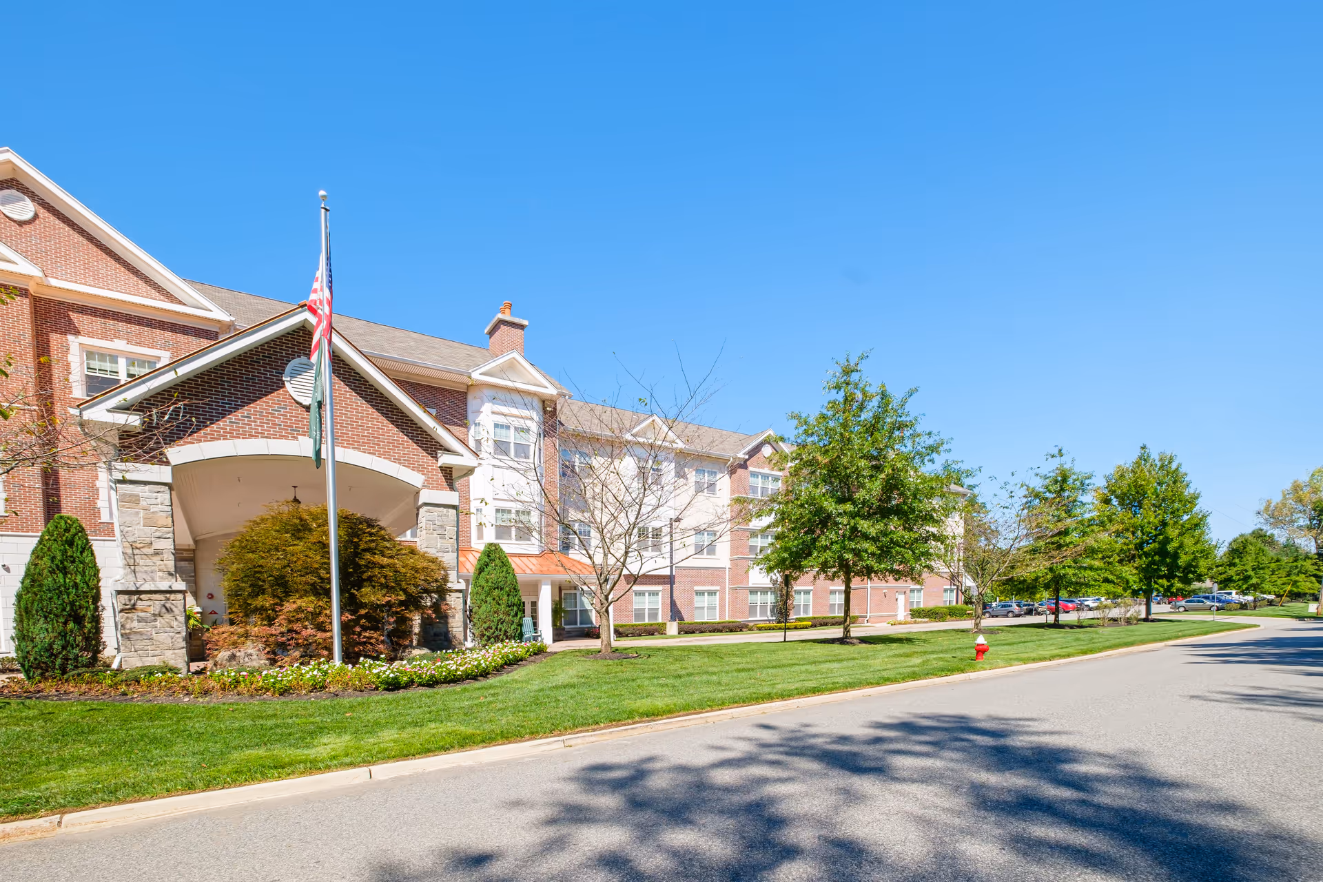 Exterior view of a senior living facility building with brick and stone facade, multiple windows, a covered entrance with an American flag, well-maintained green lawn, trees, and a clear blue sky.