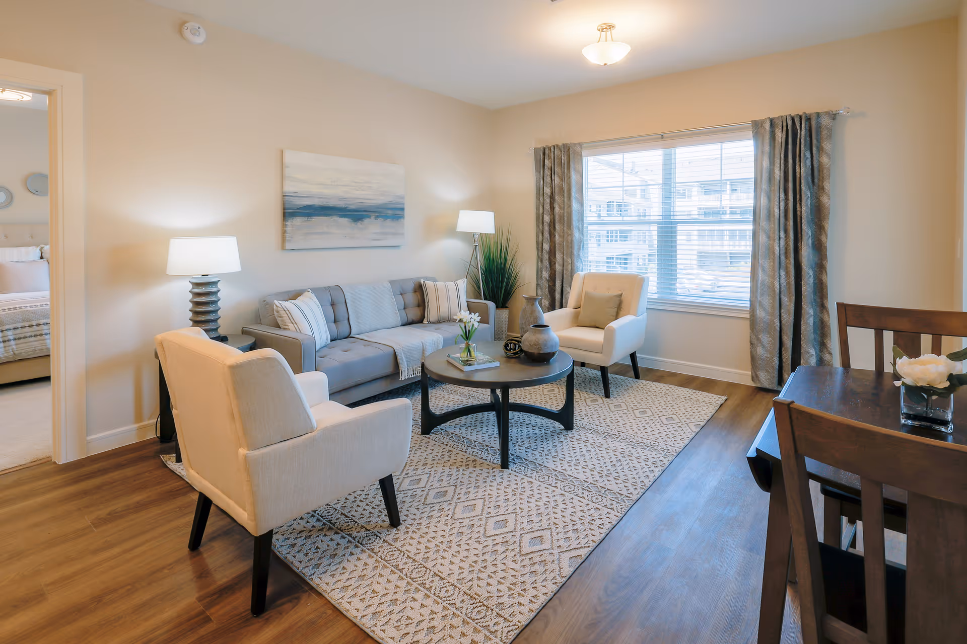A bright and cozy living room with a gray sofa, two beige armchairs, a round coffee table with decorative items, a patterned rug, and two lamps. A large window with curtains lets in natural light. A dining table with chairs is partially visible on the right, and a bedroom can be seen through an open doorway on the left.