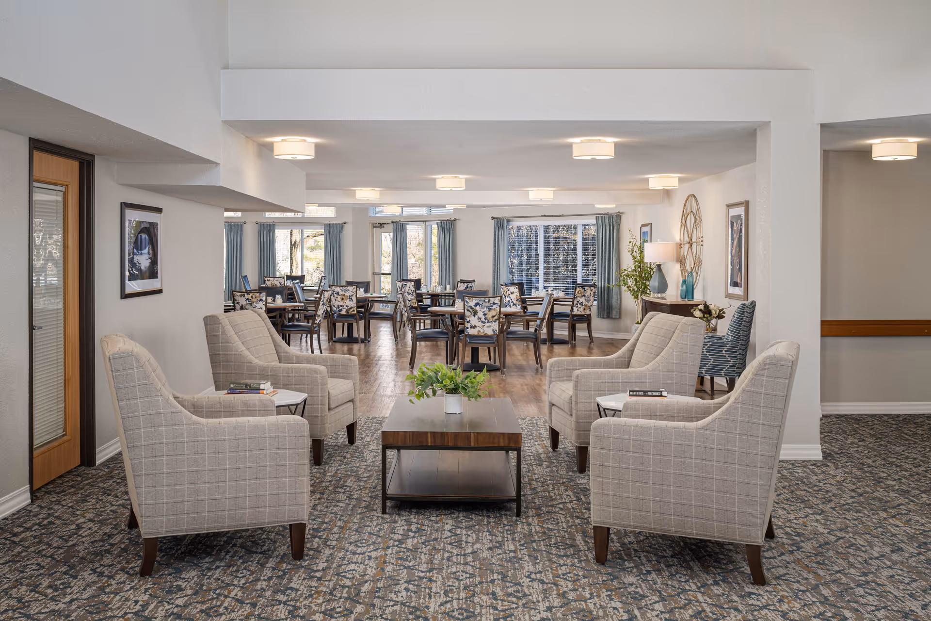 A spacious senior living common area with four beige plaid armchairs arranged around a wooden coffee table with a small plant on it. In the background, there are multiple dining tables with floral upholstered chairs near large windows with blue curtains. The room has white walls, ceiling lights, and a patterned carpet.
