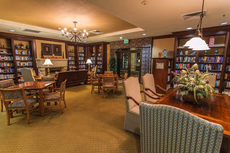 A cozy library or common room with wooden bookshelves filled with books and decorative items, several wooden tables and chairs with patterned upholstery, a fireplace with a mantle, table lamps, and a floral arrangement on a wooden table. The room has warm lighting and carpeted floors.