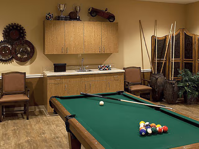 A recreation room with a green pool table in the center, pool cues resting on the table, and a rack of billiard balls arranged for a game. Behind the pool table is a wooden cabinet with a sink, trophies, and decorative items on top. Two chairs are placed on either side of the cabinet. On the right side, there are tall decorative vases with long sticks and a folding screen, along with a potted plant. The room has wooden flooring and beige walls.