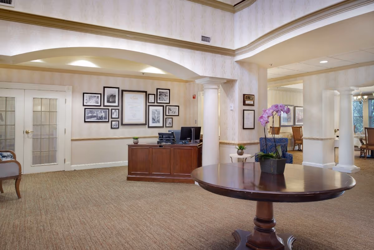 Interior view of a senior living facility lobby area with a round wooden table in the foreground holding a potted orchid. In the background, there is a wooden reception desk with a computer and printer, framed pictures and documents on the wall, and a seating area with chairs. The space features beige walls, carpeted floors, white columns, and soft lighting.
