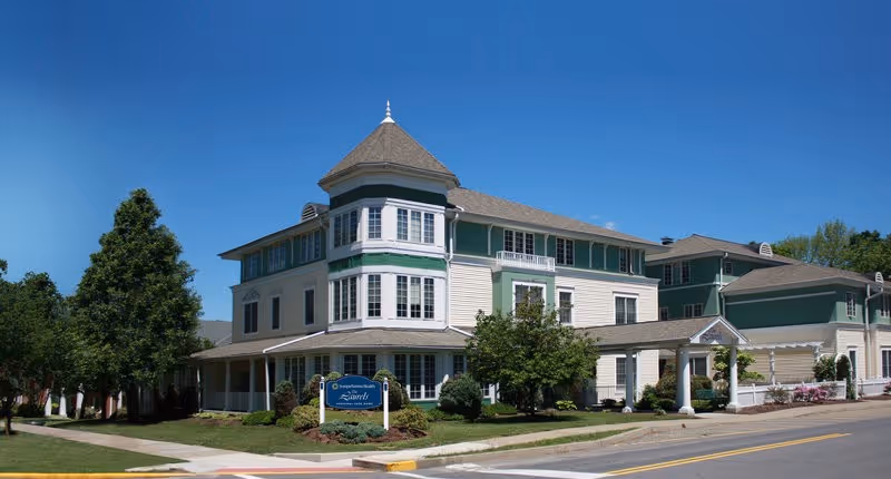 Green-and-white multi-story senior living building with a tower-like corner, landscaped grounds, and an entrance sign for The Laurels.