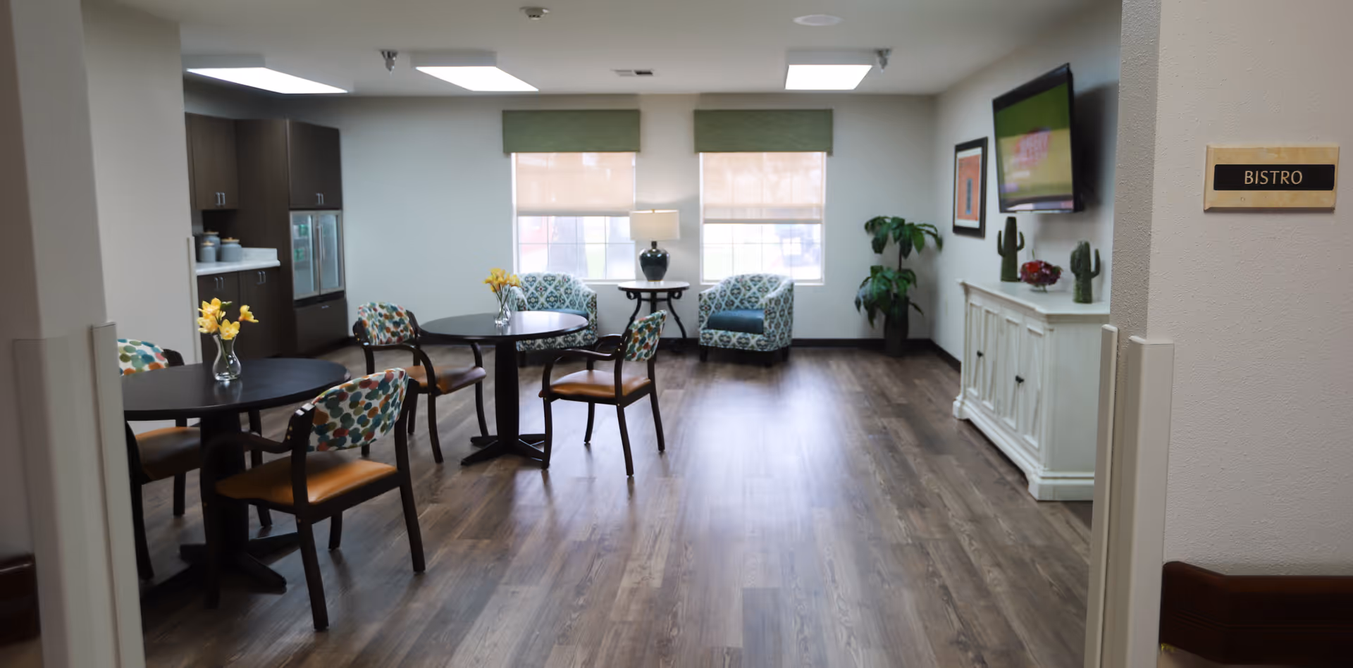 Interior view of a bistro area in a nursing and rehabilitation center with round tables and chairs, two armchairs near windows, a sideboard with decorative items, and a wall-mounted TV.