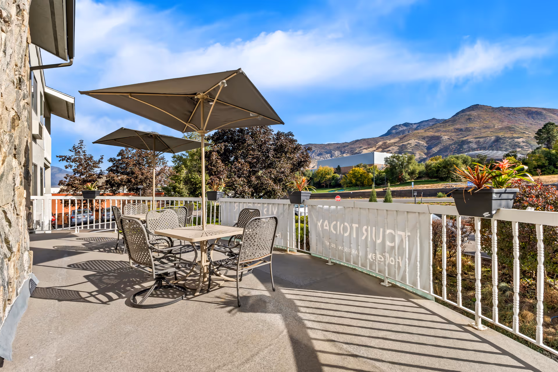 Outdoor patio area with metal table and chairs under large umbrellas, surrounded by a white railing with potted plants. In the background, there are trees, a road, and mountains under a blue sky with some clouds.