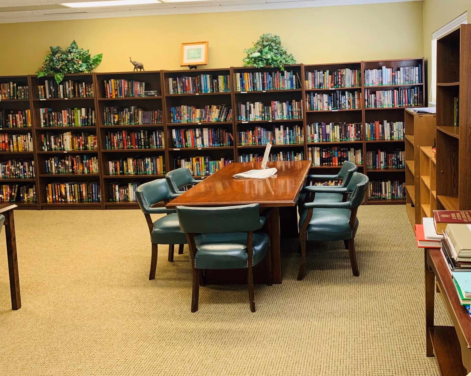 A quiet library/reading room with bookshelves along the walls and a wooden table surrounded by green upholstered chairs.