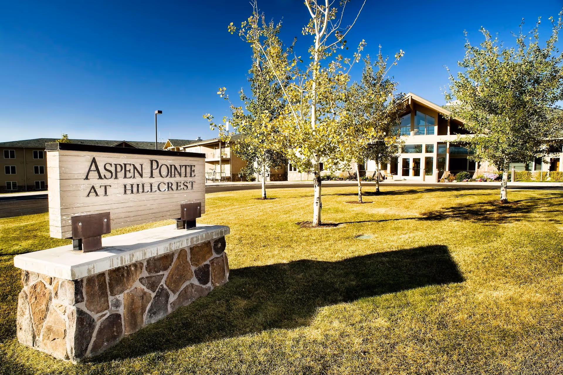 Outdoor view of Bozeman Health Hillcrest Senior Living, Aspen Pointe facility with a stone and wood sign reading 'Aspen Pointe at Hillcrest' in the foreground, green grass, several trees, and the building with large windows in the background under a clear blue sky.