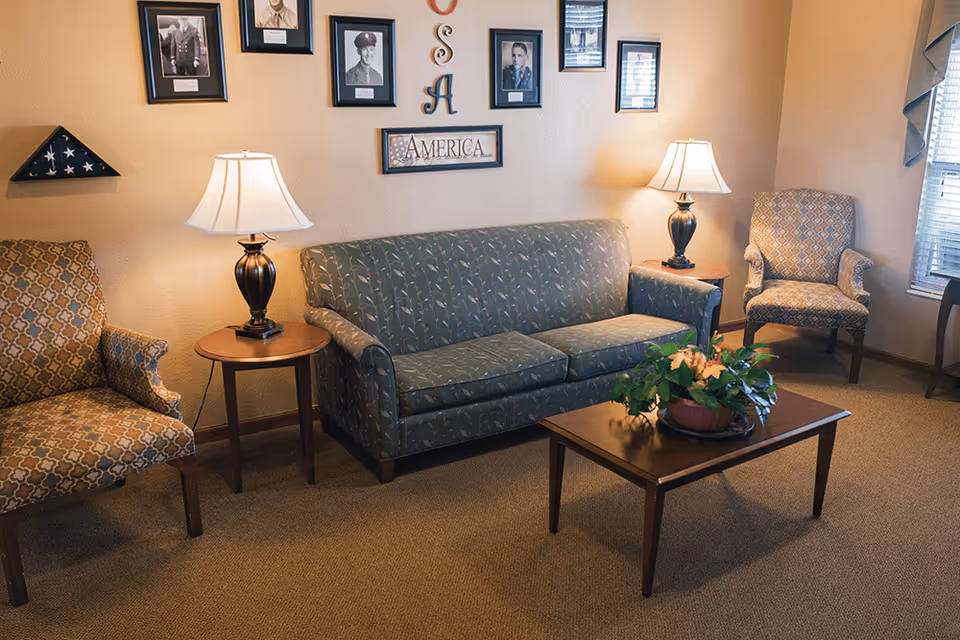 A cozy living room with a patterned sofa and two armchairs, two table lamps on wooden side tables, a wooden coffee table with a potted plant, and framed photographs and patriotic decorations on the wall.