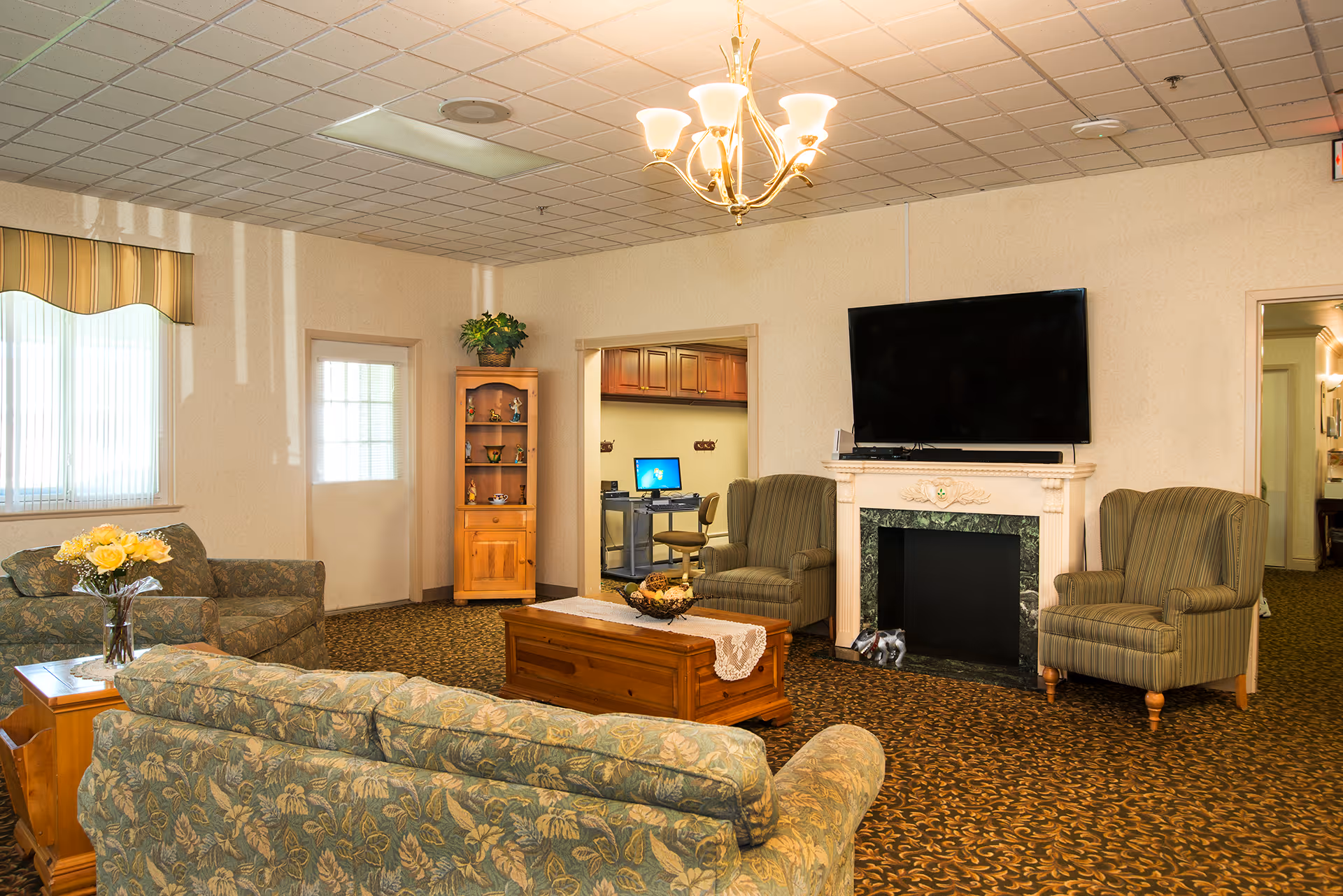 A cozy living room in a senior living facility with floral patterned sofas, two striped armchairs, a wooden coffee table with a decorative centerpiece, a flat-screen TV mounted above a fireplace, a wooden cabinet with plants and decorations, and a computer workstation visible through an open doorway.
