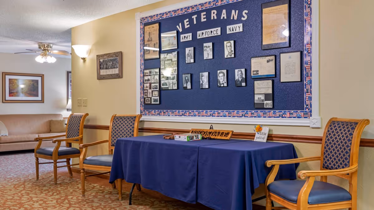 A seating area in a senior living facility with three wooden chairs with blue patterned cushions around a table covered with a blue tablecloth. Behind the table is a bulletin board dedicated to veterans, displaying photos and documents related to the Army, Air Force, and Navy. In the background, there is a beige couch and framed artwork on the wall.