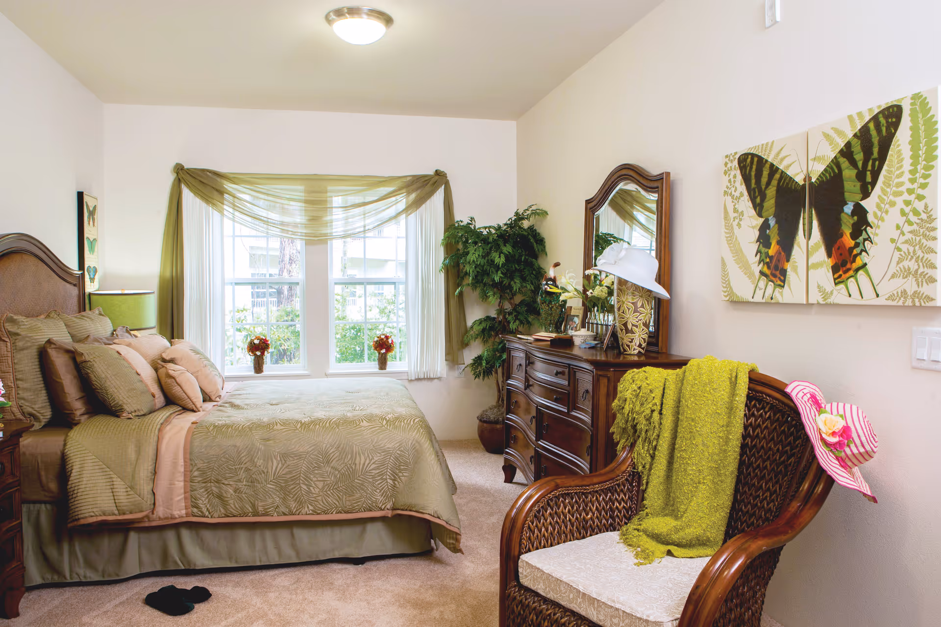 A cozy bedroom with a large bed covered in green and beige bedding, multiple pillows, and a wooden headboard. There is a window with sheer green curtains letting in natural light. A wooden dresser with a mirror and decorative items sits against the wall, next to a potted plant. A wicker chair with a green throw blanket and a pink striped hat is positioned in the foreground. Butterfly artwork hangs on the wall above the chair.