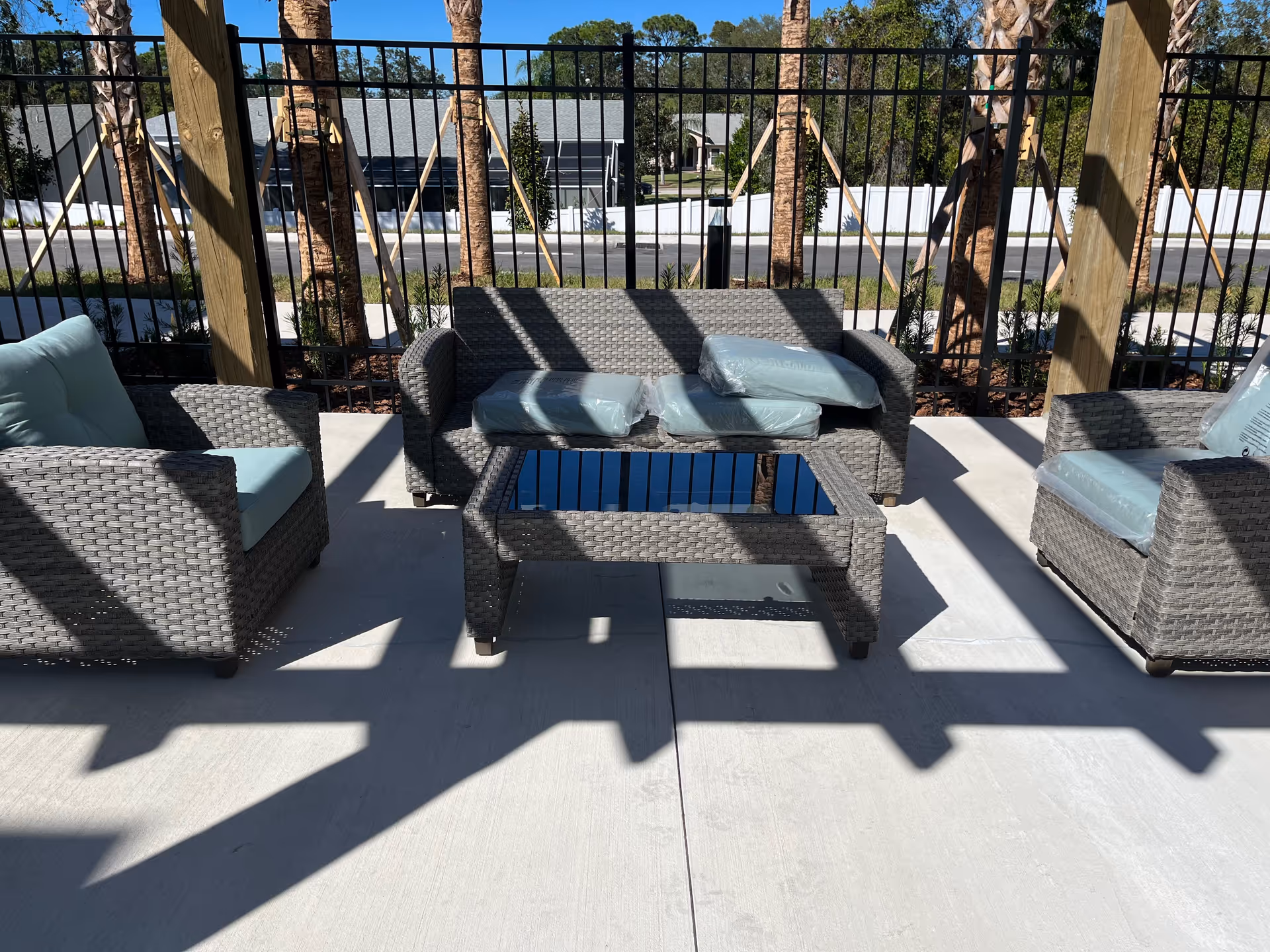 Outdoor patio area with three gray wicker chairs and a matching loveseat with light blue cushions still in plastic covers, arranged around a glass-top wicker coffee table. The patio is shaded by a wooden pergola casting shadows on the concrete floor, with a black metal fence and palm trees in the background.