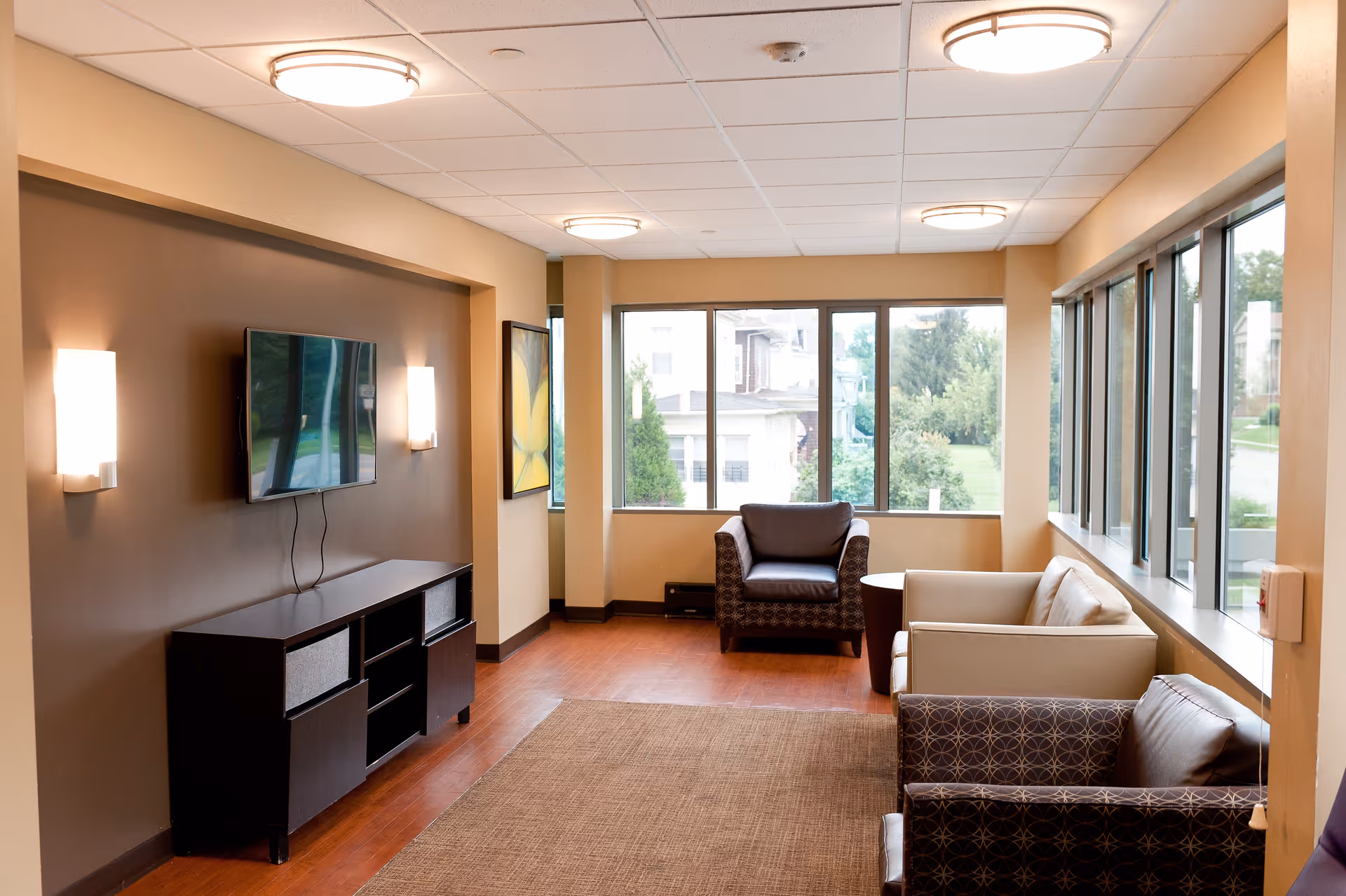 A cozy living room area with a flat-screen TV mounted on a brown accent wall, two wall-mounted lights, a dark wood TV stand, and several chairs and a sofa arranged around a small table. Large windows on two sides let in natural light and offer a view of greenery outside.