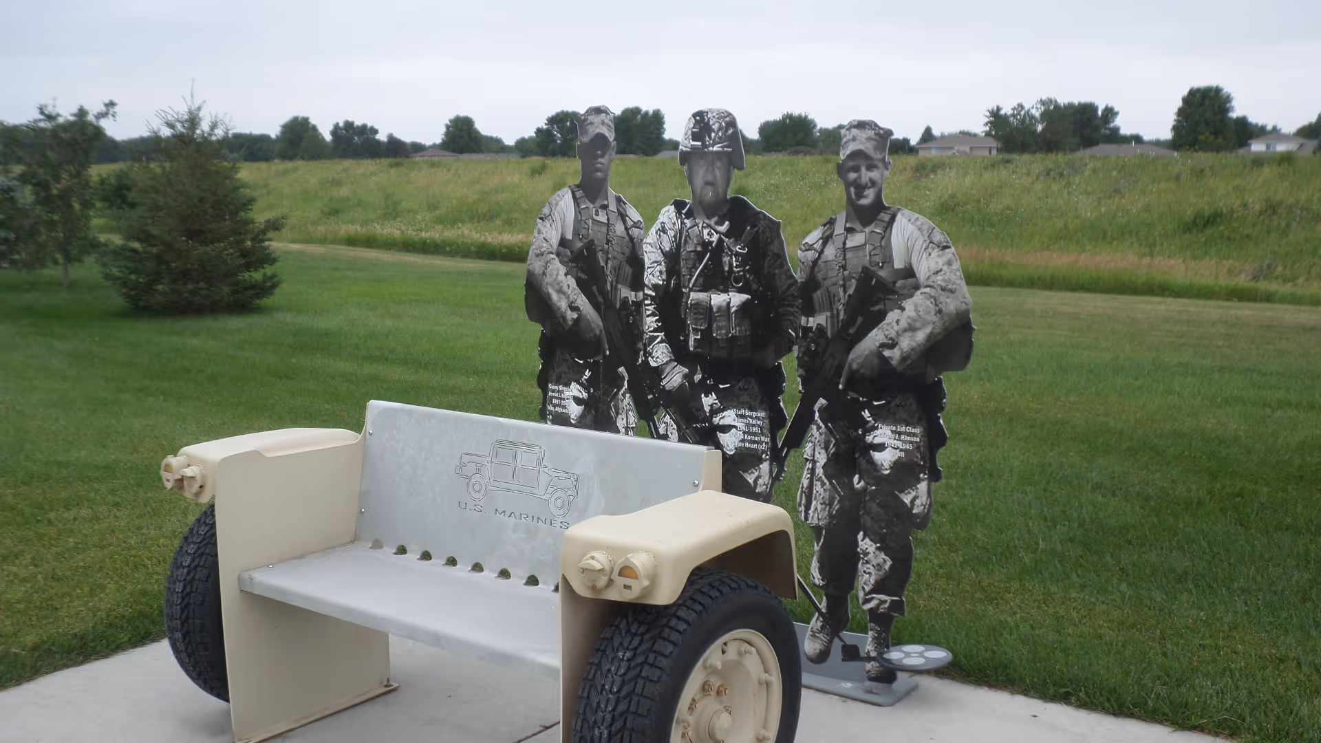 A vehicle-style bench on a concrete pad with three life-size black-and-white soldier cutouts standing behind it on a grassy field.