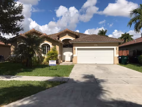 Single-story house with a tiled roof, beige exterior walls, a two-car garage, and a driveway. The front yard has green grass, a palm tree, and other plants. The sky is partly cloudy with blue patches.