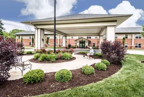 Charter Senior Living of Godfrey entrance with canopy, landscaped circle, and a resident with a dog.
