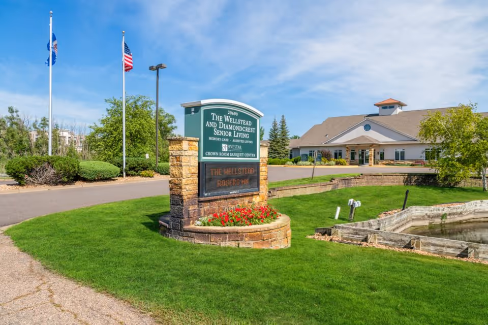 Exterior view of The Wellstead and Diamondcrest Senior Living facility with a large sign in the foreground displaying the facility name and services. The building is set behind a well-maintained lawn with a small pond and flower bed. Two flagpoles with flags are visible to the left, and the sky is clear with some clouds.