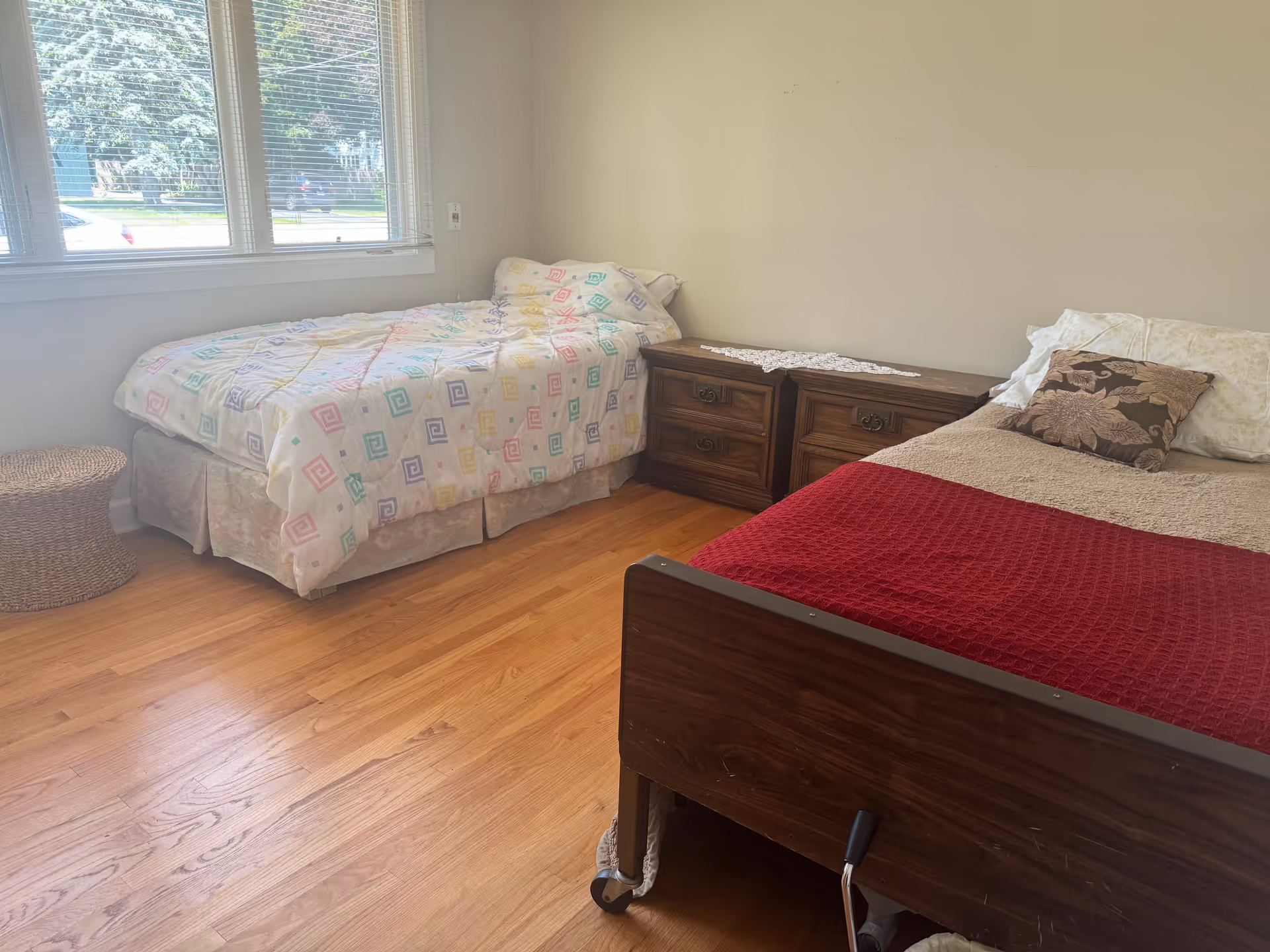 Two beds in a bright bedroom with wooden floor, dressers, and a window view.