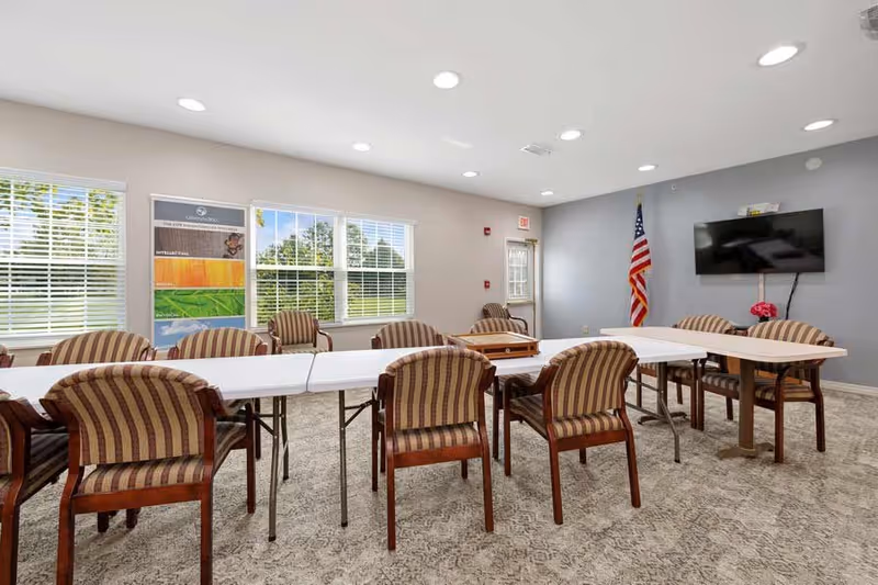 A bright meeting or activity room with several striped upholstered chairs arranged around white folding tables. The room has large windows with white blinds letting in natural light, a wall-mounted flat-screen TV, an American flag in the corner, and a colorful poster on the wall. The carpet is patterned and the walls are painted in light neutral tones.