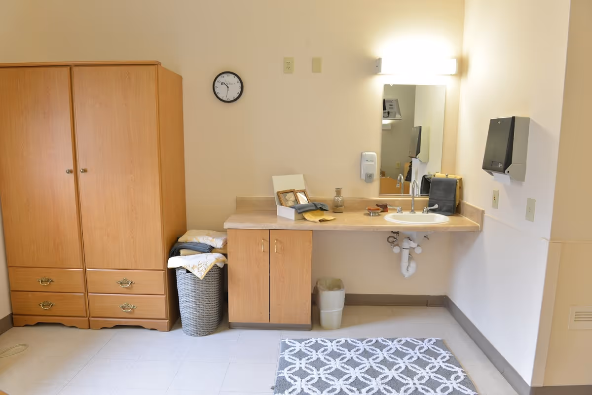 A senior living room area with a wooden wardrobe, countertop sink and mirror, storage cabinet, laundry basket, and a patterned rug.