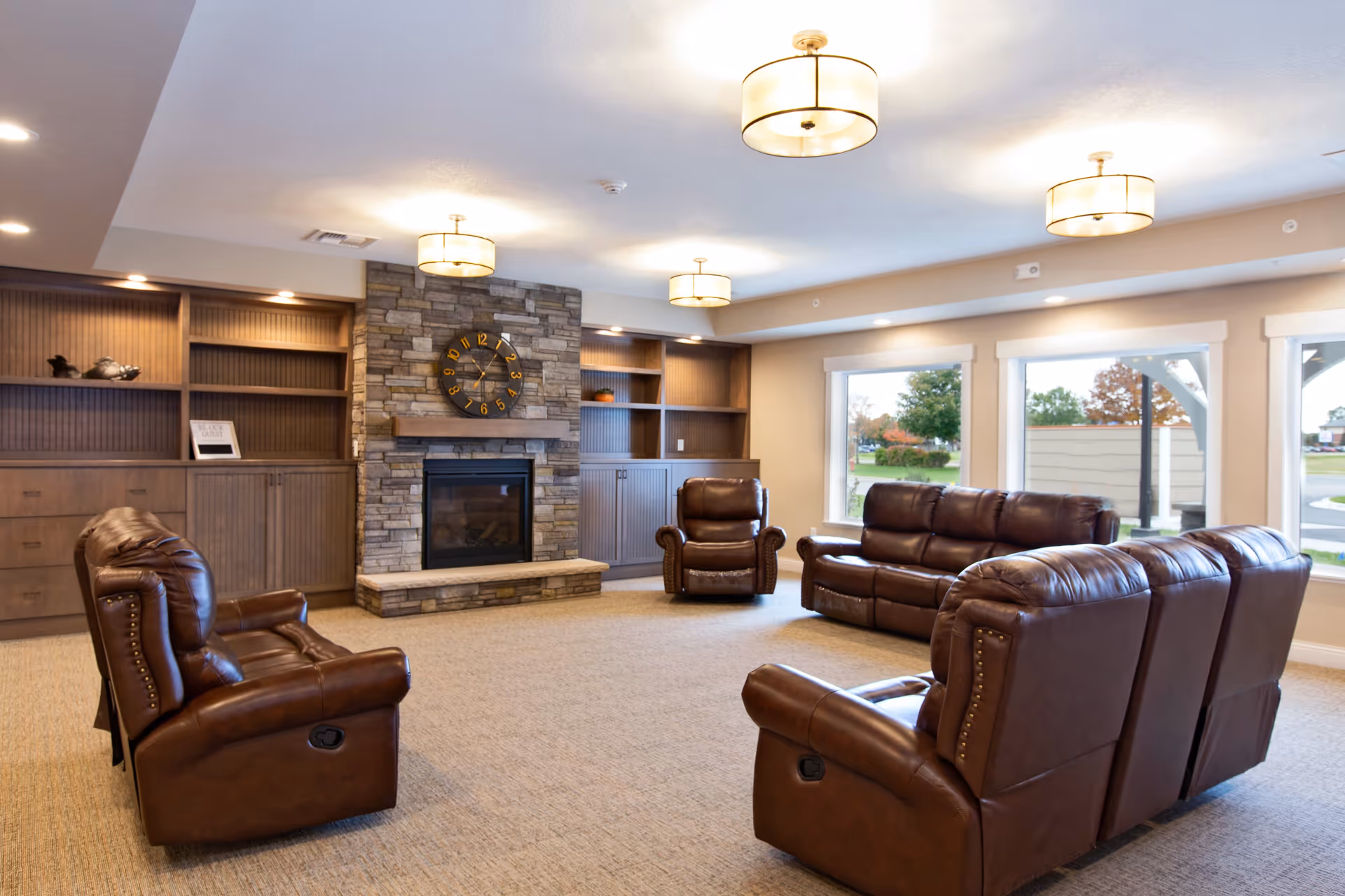A spacious living room with brown leather recliner chairs and sofas arranged around a stone fireplace with a large clock mounted above it. Built-in wooden shelves and cabinets flank the fireplace. Large windows on one side let in natural light and offer a view of the outdoors. The ceiling has multiple round light fixtures.