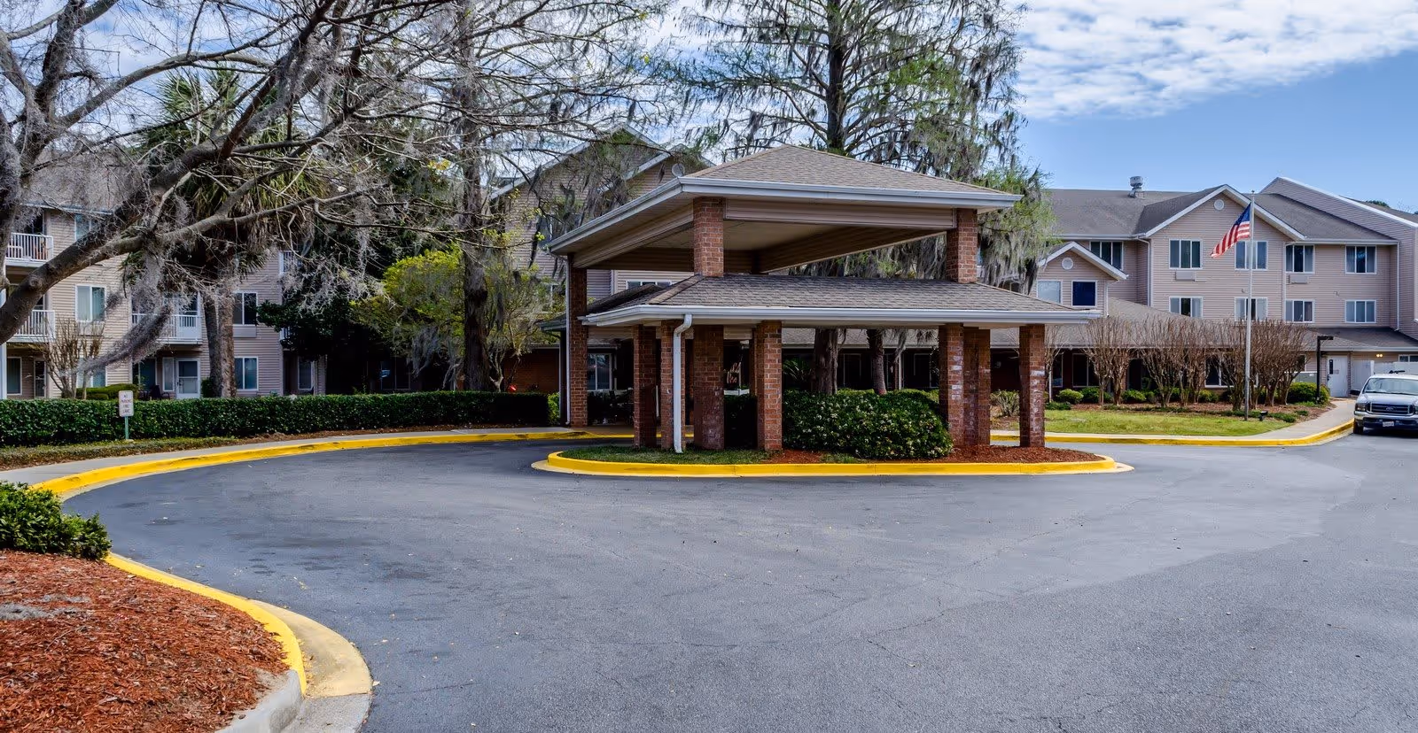 Entrance driveway of a senior living facility with a covered drop-off area supported by brick pillars. Surrounding the driveway are landscaped areas with bushes and trees, and in the background, there are multi-story residential buildings with beige siding and an American flag on a flagpole.