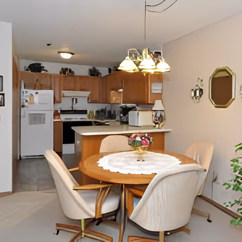 Interior view of a senior living facility dining area and kitchen. The dining area features a round wooden table with a white lace tablecloth and a centerpiece of flowers, surrounded by four cushioned chairs on casters. Above the table hangs a chandelier with multiple lights. The kitchen in the background has wooden cabinets, a white refrigerator, a stove, and a countertop with various kitchen items. The walls are light-colored, and there is a decorative mirror and wall sconces on the right wall.