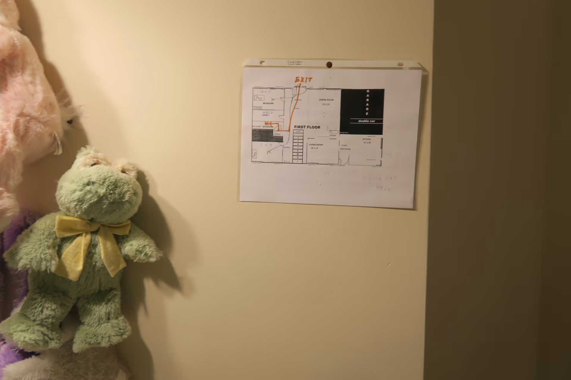 A wall with a floor plan taped to it, showing the layout of the first floor including rooms like bedroom, living room, kitchen, and garage. To the left of the floor plan, there are plush toys hanging on the wall, including a green frog with a yellow bow.
